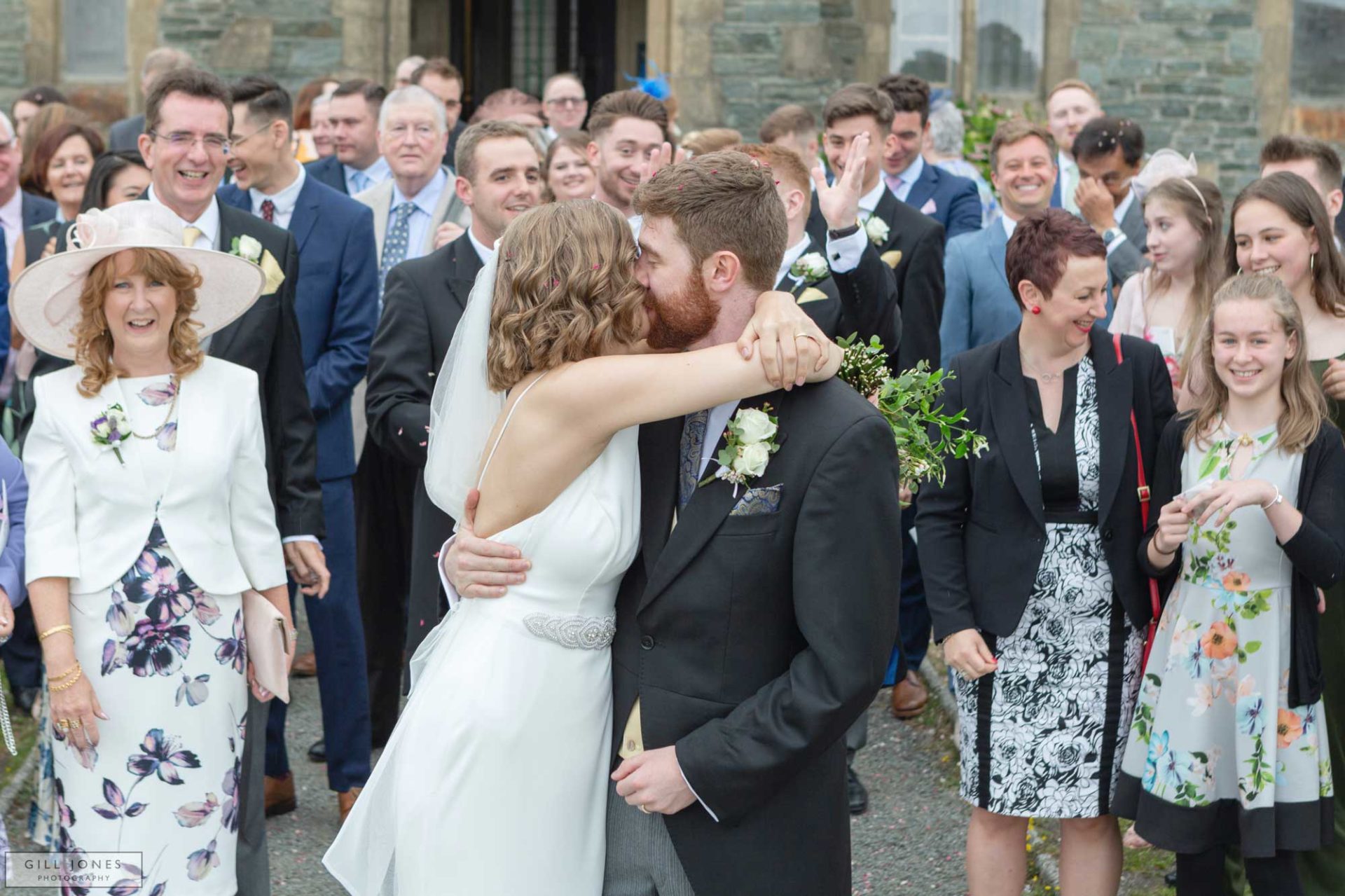 the bride and grrom are kissing each other infront of their guests outside St. Ffraids church in Trearddur Bay