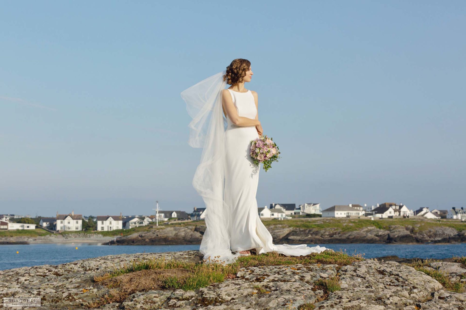 a bride standing on the rocks in trearddur bay the wind is blowing her veil
