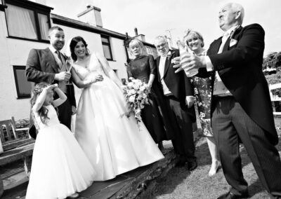 The Father of the bride opens a bottle of champagne as the guests react to the bubbles by Anglesey photographer Gill Jones Photography