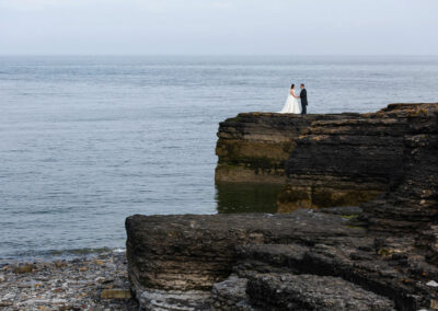 the bride and groom face each other and hold hands standing on a cliff top in Moelfre with the sea in the background by Anglesey photographer Gill Jones Photography