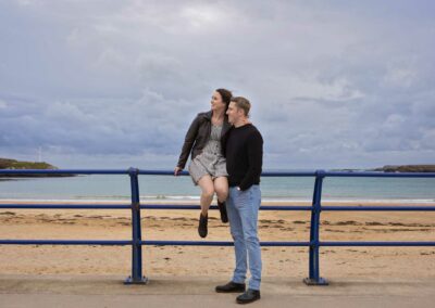 a woman sits on the top of rails at the beach as her boyfriend holds on to her by Anglesey wedding photographer Gill Jones Photography
