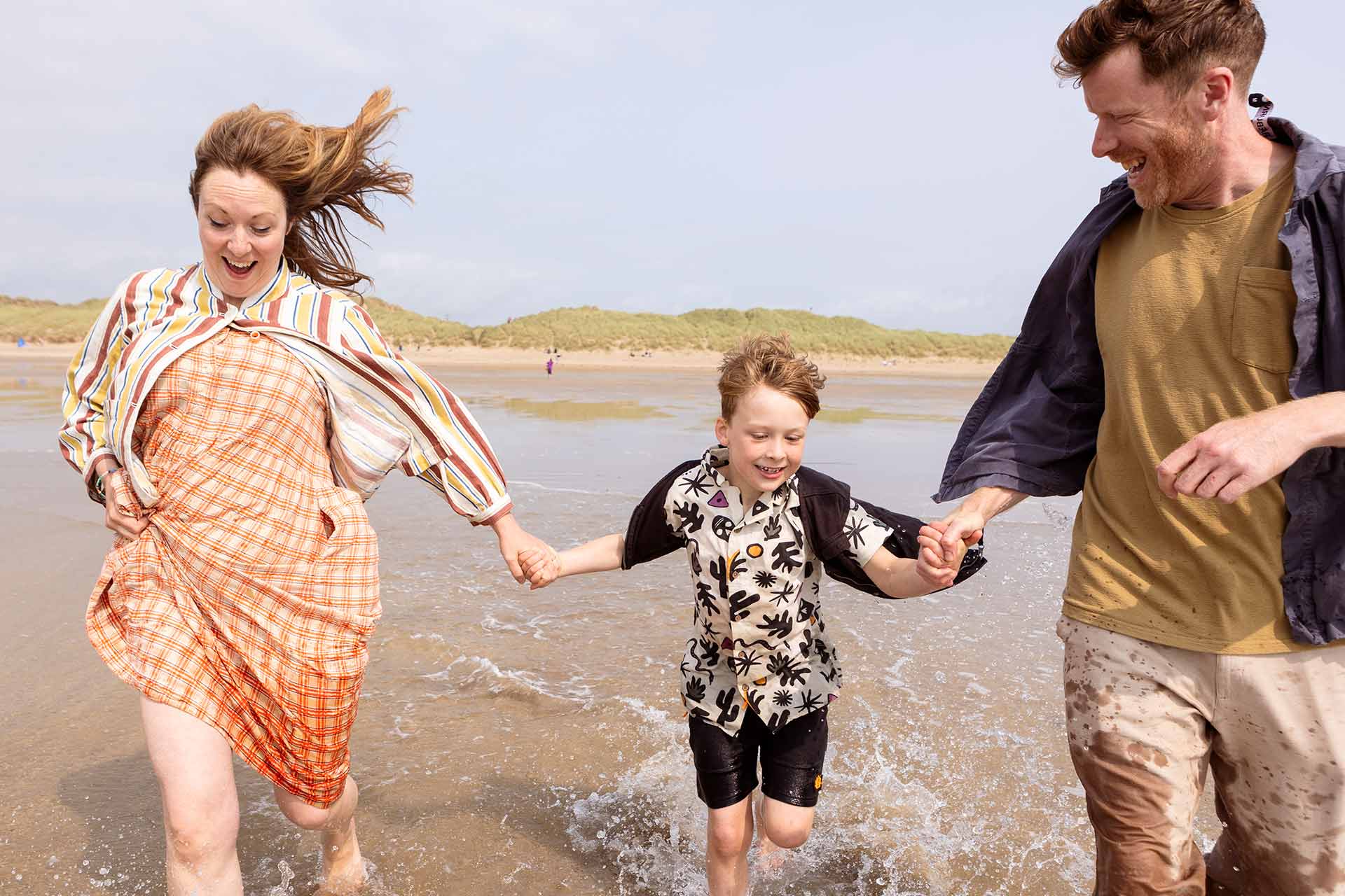 A mother and father hold hands with their son as they run into the waves at a sandy beach by Anglesey family photographer Gill Jones Photography