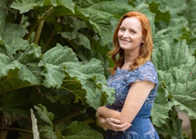 a woman wearing a blue dress, blue eyes and red hair stands with her arms crossed infront of a large leafed plant by Anglesey elopement photographer Gill Jones Photography