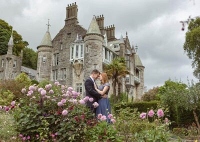 a husband and wife are located infront of a beautiful chateau they hug behind pink dahlias by Anglesey elopement photographer Gill Jones Photography