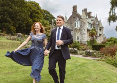 a husband and wife walk hand in hand, she swings the hem of her dress infront of a beautiful chateau by Anglesey elopement photographer Gill Jones Photography