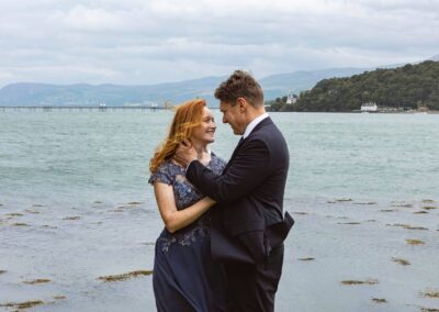 a husband and wife caress whilst standing infront of a stretch of water by Anglesey elopement photographer Gill Jones Photography