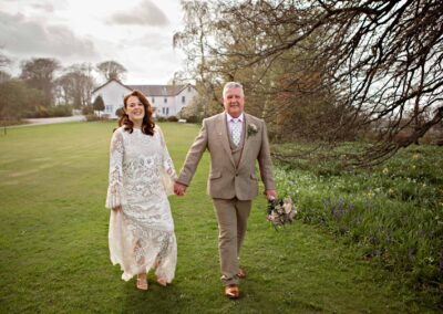 a mature bride and groom walk hand in hand with Plas Dinas Country House in the background thr groom is holding his bride's bouquet by Anglesey photographer Gill Jones Photography