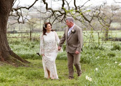 mature bride and groom walk hand in hand amongst the daffodils as they chat the bride holds her dress by Anglesey photographer Gill Jones Photography