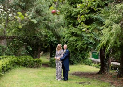 a woman stands with her back towards her husband's chest surrounded by trees, there's an apple hanging in the foreground