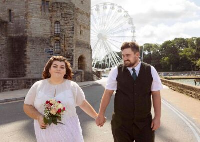 a bride and groom walk hand in hand with a castle and a fair ground wheel in the background