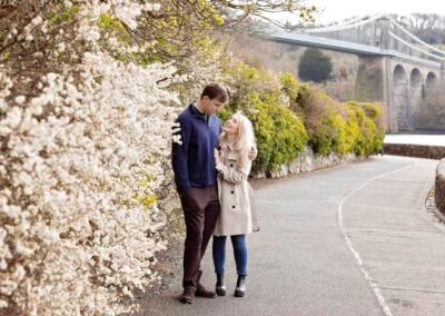 a man and woman hug as they stand by a hedge in blossom with a bridge in the background