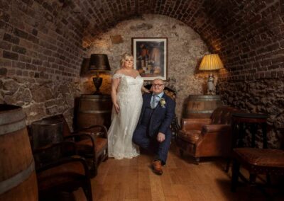 a man sits on a leather chair as his bride stands by his side they're in a wine cavern