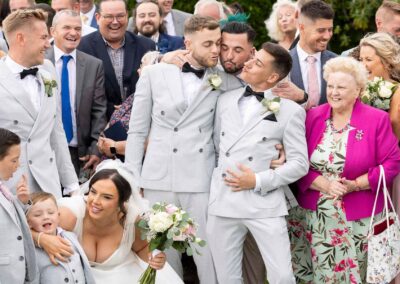 a group of wedding guests gather round whilst three groomsmen lean together to kiss one another