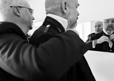 a groom reaches his arms over his brother's shoulders to tie a neck tie