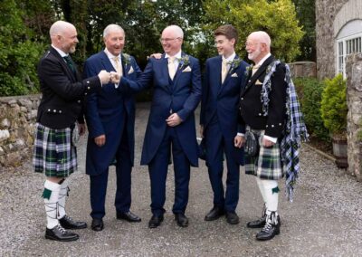 a groom, surrounded by his friends, fist bumps a man wearing a kilt.