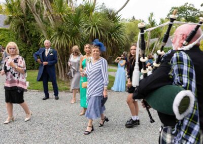 a group of wedding guests walk past and acknowledge a man playing the bagpipes