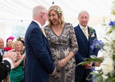 a bride faces her husband to be at the alter and they laugh together, her father looks on